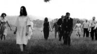A gaggle of ghouls, men and women among them, looking disheveled and bedraggled, lurch across a field.