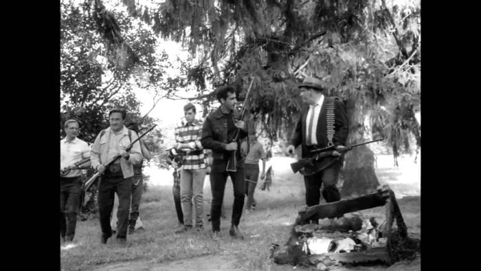 A group of men, all armed with rifles, make their way through a field, past trees and burnt debris strewn on the ground.