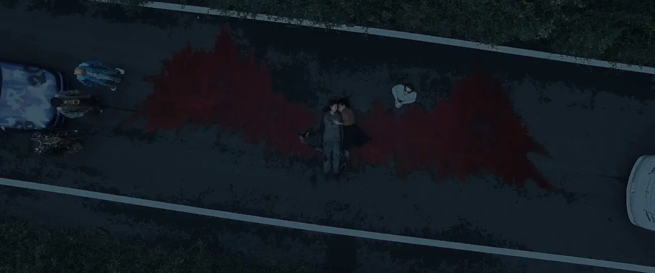 three men watch beside their car as aman lays in the middle of the road. The pattern of blood on the road looks like red wings. Another man with glasses on looks up towards the sky.