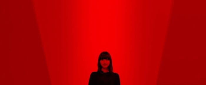 In an all red room with with red lighting a long-haired girl smiles at the camera 