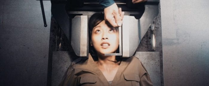 A girl lying on a metal slab and her head between two clamps looks anxiously at a hand hoovering above her, measuring her face.