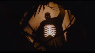 A shadow of a man can be seen through a hole shrouded in darkness. The man's rib cage is bright white and there is light in the shadow where his eyes should be.
