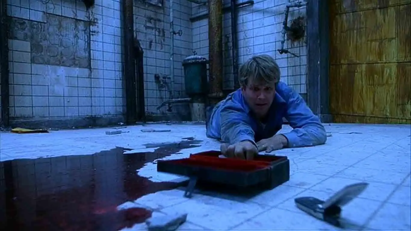 Lawrence Gordon reaching for a cell phone with a pool of blood covering the floor underneath him.