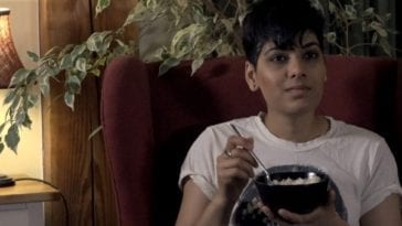 a woman sits in a padded chair in a well lit room smiling at the camera while eating a bowl of rice.