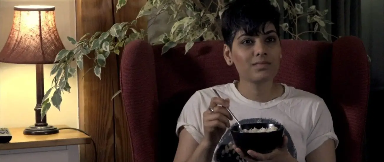 a woman sits in a padded chair in a well lit room smiling at the camera while eating a bowl of rice.