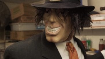 A man wearing a suit, a pilgrim hat and a grinning with unkempt hair wields a hatchet in his gloved left hand while standing in a restaurant kitchen