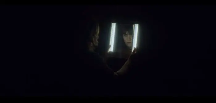 Catherine stares into a brightly lit mirror while the mirrored face stares back toward the camera.