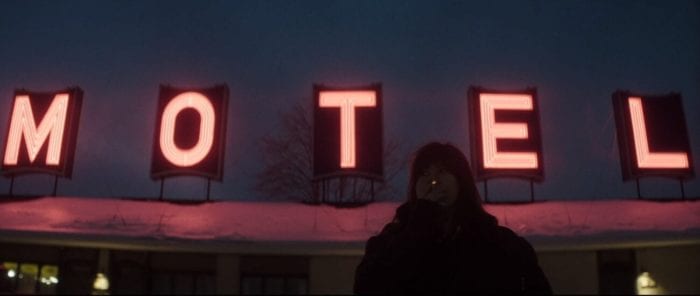 Catherine stands outside in a heavy winter jacket smoking a cigarette. A neon sign reads "MOTEL" on the roof's overhang above her.