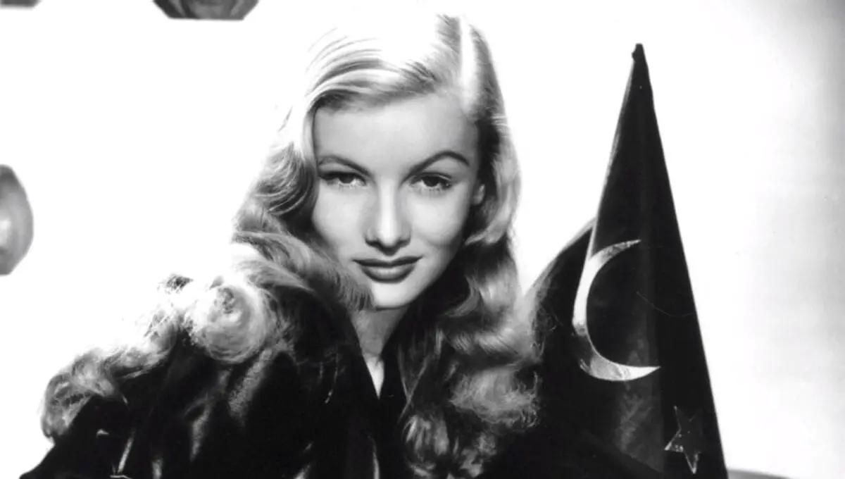 Veronica Lake stares into the camera over her right shoulder against a white backdrop with a witch's hat adorning a half moon on it.