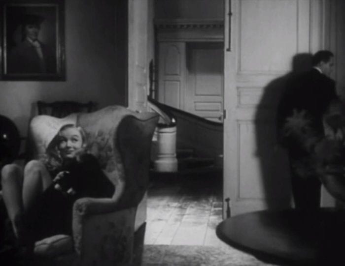 Veronica Lake sits in a chair smiling and petting her black cat while Frederic March investigates the room's light switch