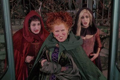 The three Sanderson sisters stand in front of open bus doors.