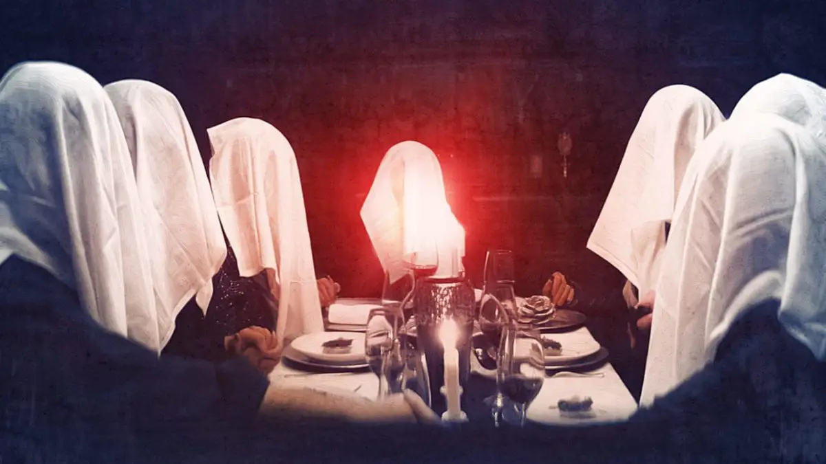 The Sinclairs sit at a dining room table holding hands with their napkins over their head and an orb of red light floating from the center of the feast