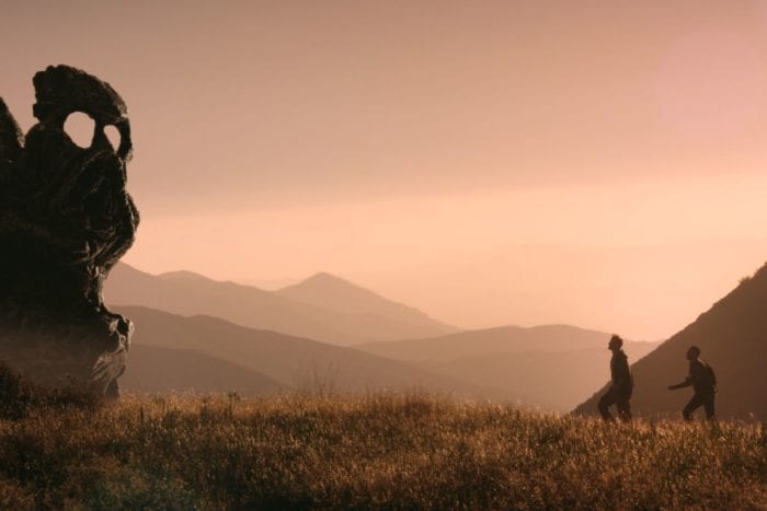 Two men walking towards a large stone statue in a grassy mountain range at sunset.