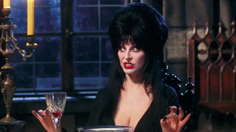 Cassandra Peterson as Elvira, Mistress of the Dark, sits at a dinner table, unhappy with what's going on.