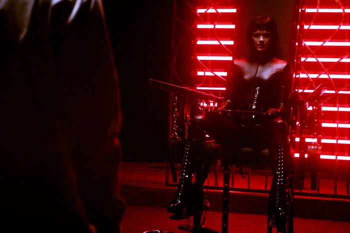 A pale woman with a black bob-style haircut dressed head-to-toe in leather is sitting on a chair in front of red neon lights in an otherwise dark room.