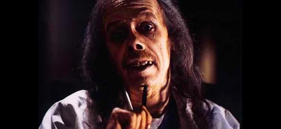 John Carpenter as the undead host of Body Bags; his skin is a sickly greenish color, his teeth are crooked and rotted, he has dark circles under his eyes, and his hair is long and stringy.