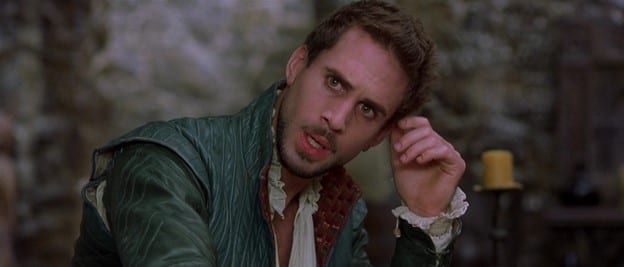 Shakespeare, as played by Joseph Fiennes, showing the beginnings of a goatee, looks up, perturbed, from his writing; he stares fixedly at something offscreen, with his left arm bent at the elbow, hand nearly touching the side of his head, which is slightly cocked to one side; a stone wall and a candle are visible in the background.