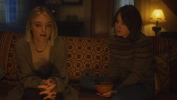 Emily (Lauren Beatty) and Rowan (Lee Marshall) sit on a couch, having an intimate conversation by the light of the fireplace.