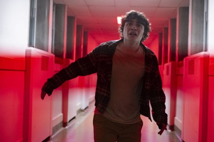 A pale boy with dark messy hair running down a white hallway lit by red light. He looks terrified.
