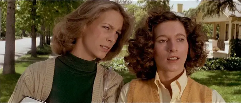 A medium shot of Laurie and Annie as they walk down the sidewalk with trees, grass and houses visible behind them; Laurie, with long, straight, light brown hair and a book just visible in her arms, has her head tilted slightly toward Annie and her eyes are slightly squinted; Annie, with long, curly, dark brown hair is shoulder to shoulder with Laurie; both girls appear to be looking at (or for) something ahead of them offscreen.