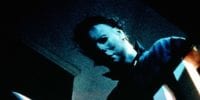 Michael Myers, half his pale white mask covered in shadow, peers over the railing at the top of the stairs; just visible at the bottom of the frame is the tip of the knife he holds in his right hand.
