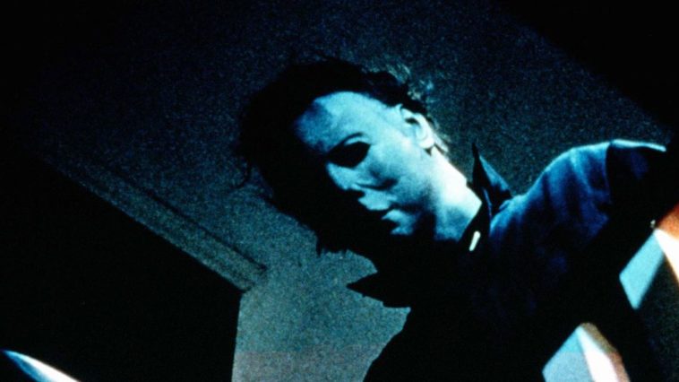 Michael Myers, half his pale white mask covered in shadow, peers over the railing at the top of the stairs; just visible at the bottom of the frame is the tip of the knife he holds in his right hand.