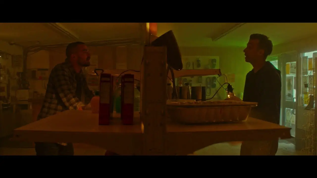 Todd, left, a man with a buzzcut wearing a plaid jacket, leaning over a barren shelf in a gas station shop lit by dim red light. Ezra, right, a man smaller than Todd with updone dark hair wearing a dark hoodie, talking to Todd on the other side of the shelf wall, standing in front of lime green light filtering in through the entry doors.