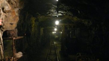 An underground mine, illuminated by overhead lights. Tools are scattered on shelves on either side of a central tunnel.