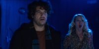 Adam Pally stands front with a shocked look on his face while Anna Camp stands behind him terrified in a dark blue room.