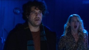 Adam Pally stands front with a shocked look on his face while Anna Camp stands behind him terrified in a dark blue room.