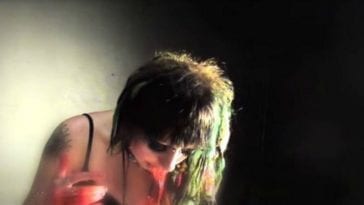 A slightly grainy image of a pale girl, her head hanging down while a red substance spills from her mouth. She has a tattoo on her right shoulder and is wearing a black tank top. Her hair, a mixture of mint green and deep brown, is partially illuminated by light coming from the right side of the image. The right side of the background is pitch black while the left is a dirt off white color, seemingly lit by the same light off to the right.