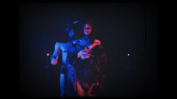 In a dark room, a woman wrapped only in a fur blanket stands in front of a shirtless man in what appears to be a bull mask. There are two candles behind them on either side of the image. The two people are lit in low red and blue lighting.