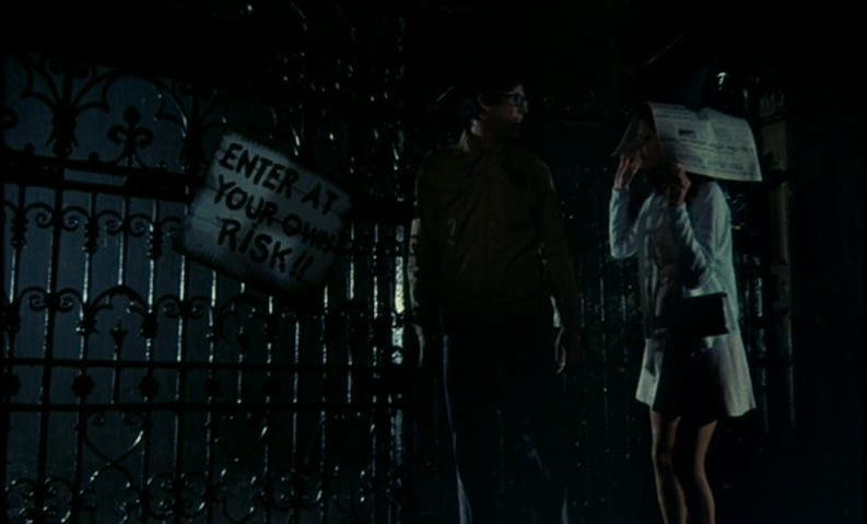 Brad Majors (Barry Bostwick) and Janet Weiss (Susan Sarandon) stand in the rain next to a sign that says, "ENTER AT YOUR OWN RISK!!", in the film, "The Rocky Horror Picture Show" (1975).