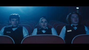 Ricky (pale boy with thick brows and short dark hair), Chaz (pale girl with heavy eyeliner and dark hair tied back in a ponytail), and Abe (husky pale boy with shoulder-length fuzzy dark hair) sit in a vacant theater watching the cursed film reel. They all wear black vests adorned with golden nametags atop white dress shirts, and black bowties. They are illuminated from behind by the soft blue projector light. They all appear concerned, Chaz especially, her mouth agape and eyes wide.