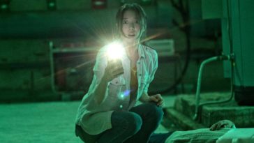 Yoo-mi shines her phones flashlight into the camera while crouching over a body in a green lit basement with a surprised action on her face
