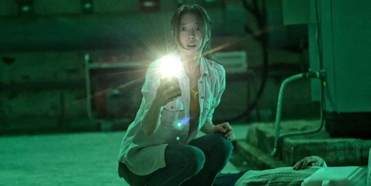 Yoo-mi shines her phones flashlight into the camera while crouching over a body in a green lit basement with a surprised action on her face