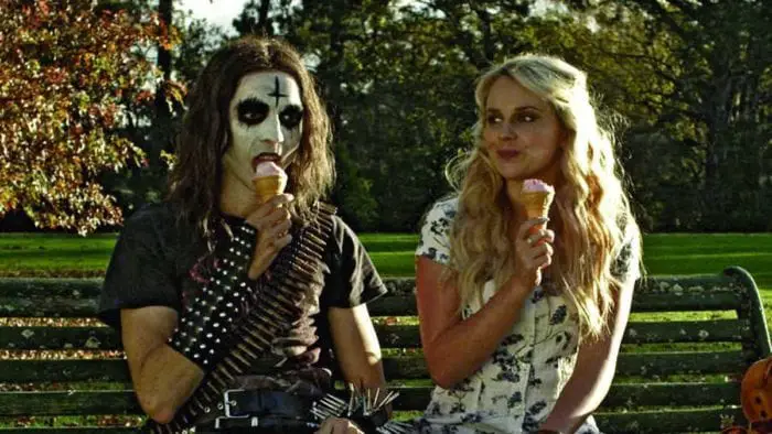 Brodie and Medina sit on a bench eating ice cream cones. Brodie is dressed in spikes and white face paint with a black upside-down cross on his forehead while Medina is in a flowery dress