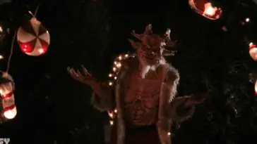 Satan Claus (Jana Peck) shrugs with a smile, wearing a Santa coat, in the film, "Letters to Satan Claus" (2020).