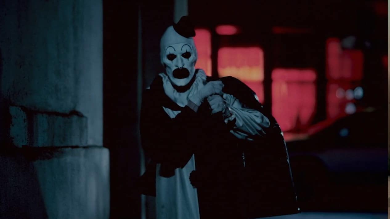Art the clown next to a building in front of another building glowing with inside red lighting. He's carrying a black trash bag and is frowning slightly. His suit is white on the right, black on the left, and the sleeves are inverted. He wears a tiny black top hat.