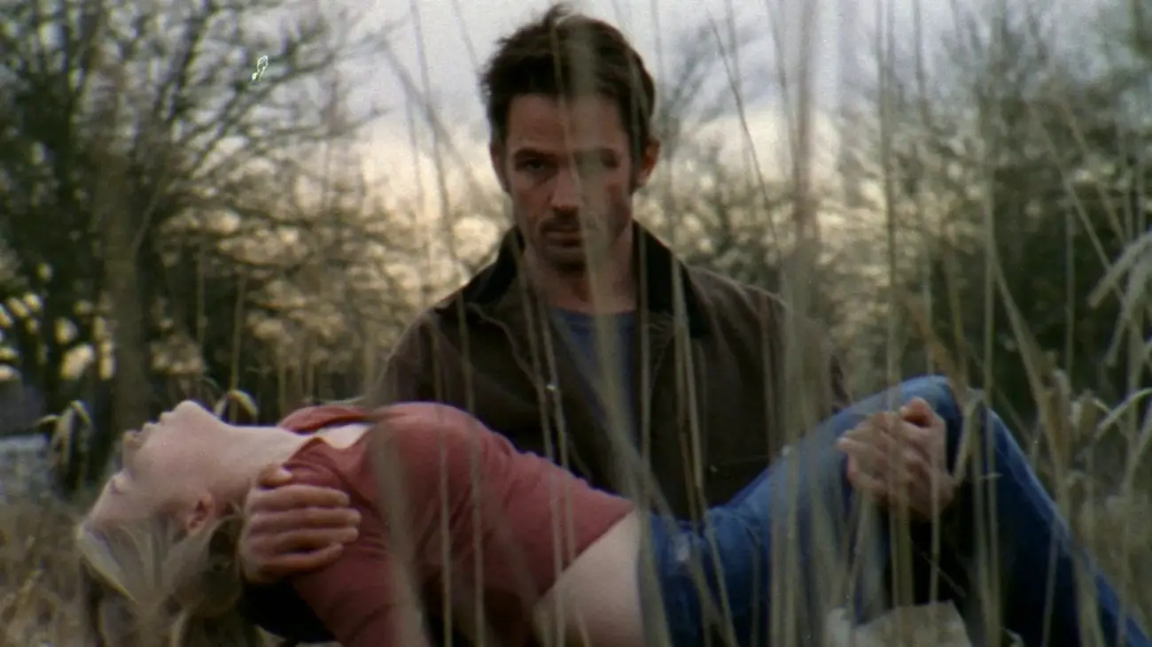 A man with a grim expression carries an unconscious woman through a field.