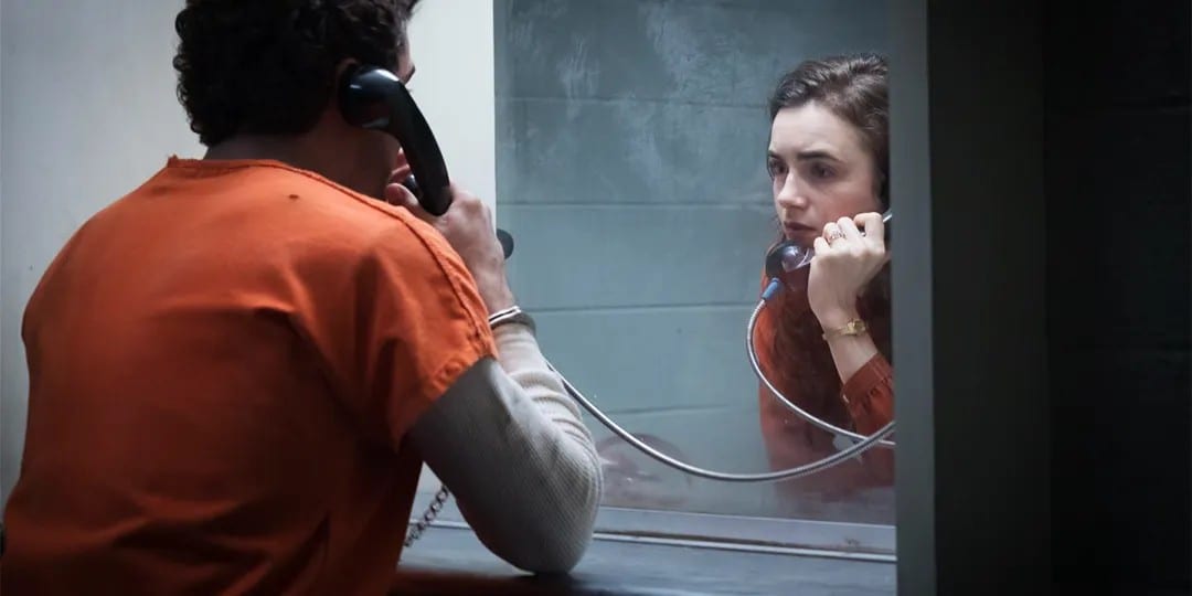 A man in an orange jail-issue jumpsuit sits with his back to the camera as he speaks to a woman on a phone receiver through a glass partition.