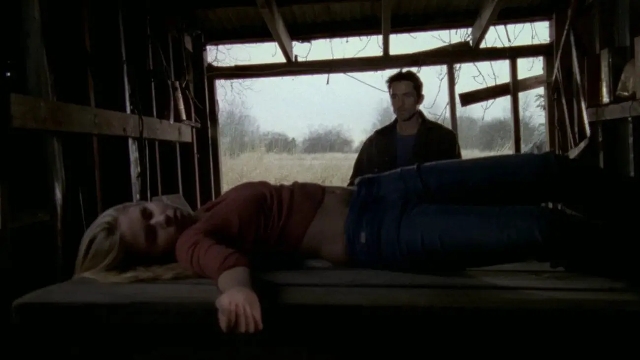 A man stands in the background looking at an unconscious woman lying on a wooden table. They stand in what looks like an abandoned barn or shed. The space behind him is open to the outdoors in an isolated, rural area.
