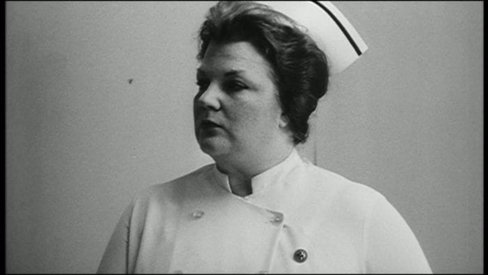 A woman in a nurse's uniform with a serious look on her face looks at something off-camera.