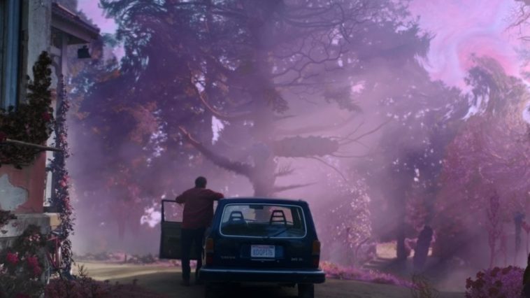 A man stands outside of his car in front of trees and looks at purple mist.