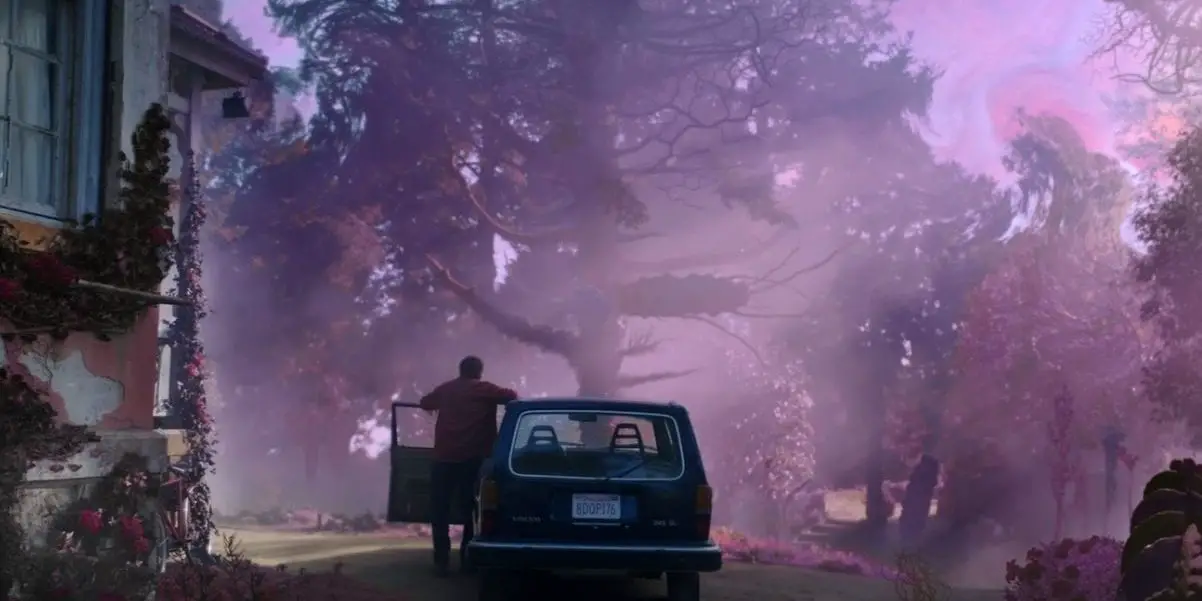 A man stands outside of his car in front of trees and looks at purple mist.