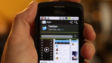A hand holds a mobile phone with the Twitter app displayed on the screen.
