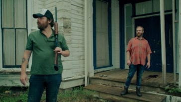 Hank and Wade stand in front of a large house. Hank is wearing a hat and carrying a shotgun.