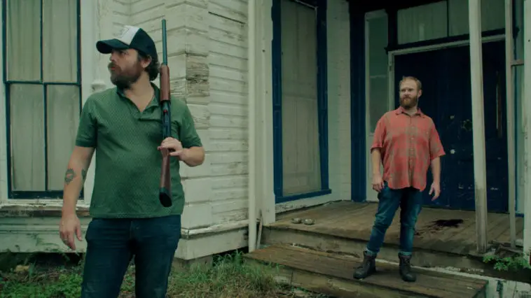 Hank and Wade stand in front of a large house. Hank is wearing a hat and carrying a shotgun.