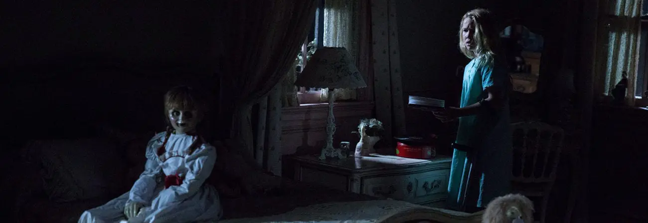 In a dark, shadowy room, a vintage doll sits on a bed. A young girl looks at the doll from the shadows.