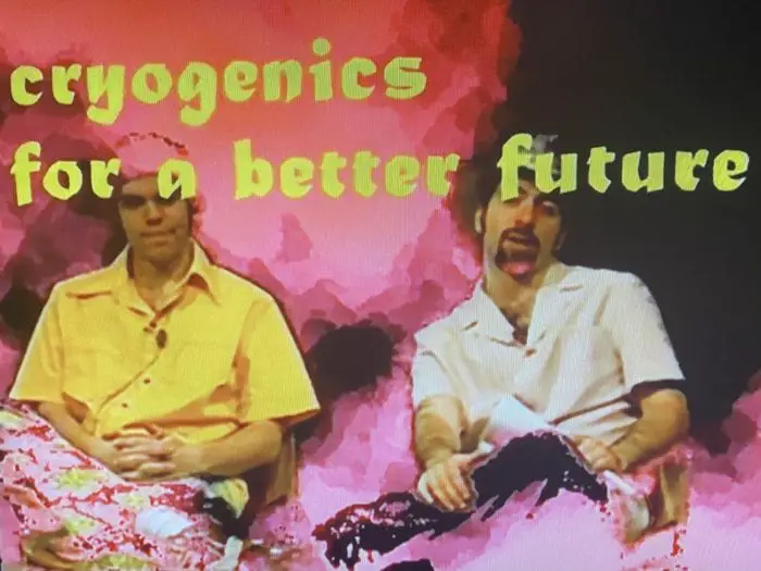 heavily filtered image of two men sitting on a tv set of a public access show - the words cryogenics for a better future superimposed on the screen